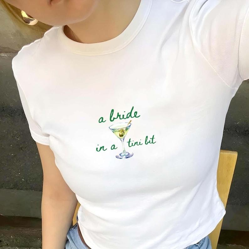 A Bride In A Tini Bit Baby Tee, Martini Bachelorette Party Shirt Hoodie, Sweatshirt | HypeCrazeTee.com